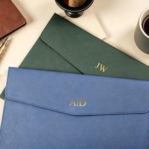 Puede incluir: Dos portadocumentos de cuero personalizados, uno azul y otro verde, con iniciales doradas. También se ven un diario de cuero marrón, un bolígrafo dorado y una taza de café. Los artículos están dispuestos sobre una superficie beige claro.