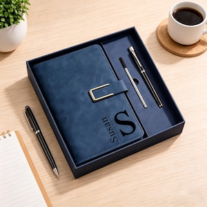 May include: A navy blue leather journal with a gold clasp, personalised with the name "Susan" and the letter "S". The journal is in a gift box with two pens. A cup of coffee and a small plant are also visible.