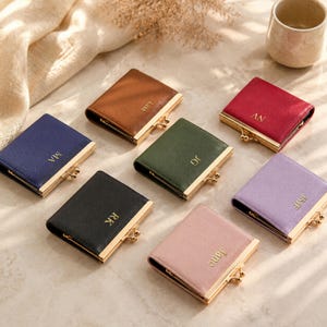 Puede incluir: Siete carteras cuadradas de varios colores, incluyendo marrón, rojo, azul oscuro, verde oliva, negro, lavanda y rosa claro. Cada cartera cuenta con un cierre de metal dorado y está personalizada con iniciales o nombres estampados en oro.
