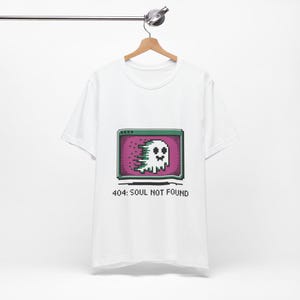 May include: A white short-sleeved t-shirt featuring a pixel art graphic of a ghost inside a computer window on a purple background. The ghost appears to be glitching with green and white pixels. Below the graphic is the text 404: SOUL NOT FOUND.