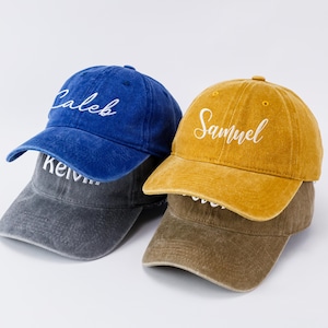 May include: Three baseball caps in blue, gray, and mustard yellow. Each cap has a curved brim and a script name embroidered on the front: "Caleb", "Kelvin", and "Samuel". The caps are stacked on top of each other.
