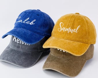 May include: Three baseball caps in blue, gray, and mustard yellow. Each cap has a curved brim and a script name embroidered on the front: "Caleb", "Kelvin", and "Samuel". The caps are stacked on top of each other.