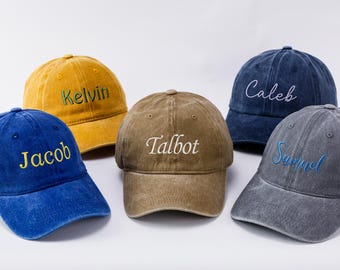 May include: Five personalized baseball caps in various colors: mustard yellow, blue, brown, navy, and gray. Each cap features a different name embroidered in a contrasting color: Kelvin, Jacob, Talbot, Caleb, and Samuel.