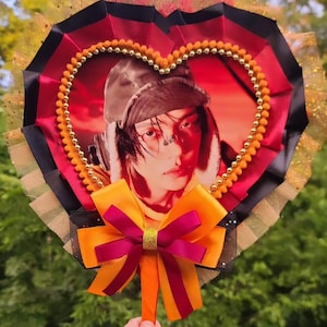 May include: A heart-shaped fan featuring a central portrait of a person wearing a brown hat against a red background. The fan is framed by red and black pleated ribbon, gold beaded trim, and a layered orange and maroon bow at the base handle.