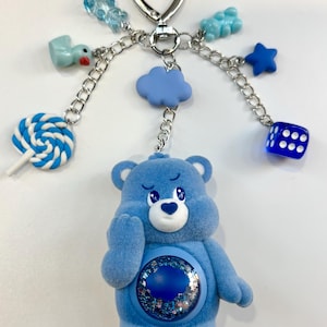 Peut inclure: Un porte-clés Care Bear bleu avec un fermoir argenté en forme de cœur. Le porte-clés comprend divers breloques, dont un nuage bleu, une étoile, un dé, un canard, un ours en gélatine et une sucette tourbillonnante bleue et blanche. L'ours a un cœur sur le ventre.