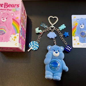 Könnte beinhalten: Ein Care Bears Colorful Hugs Grumpy Bear Schlüsselanhänger mit einer blauen Bärenfigur mit rundem Schüttelbauch, befestigt an einem herzförmigen Ring mit Anhängern wie einem blauen Lutscher, einer Ente, einer Wolke, einem Stern, einem Würfel und zwei Gummibärchen.