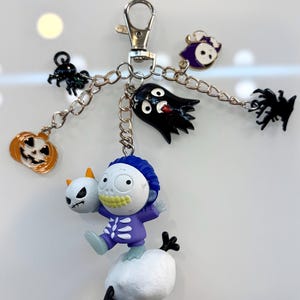 Pode incluir: Um porta-chaves de metal com uma figura central de cabelo azul num fato de esqueleto roxo que segura um pequeno crânio branco sobre uma base branca. Estão anexados berloques de gato preto, abóbora laranja, fantasma e bruxa.