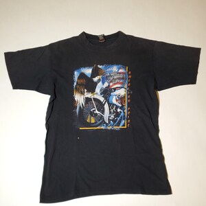 Peut inclure: T-shirt noir avec un motif de moto, d'aigle et le texte "American Pride". Le design comprend des éléments rouges, blancs et bleus. Le logo Harley-Davidson Cycles est également visible.