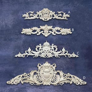 Può includere: Quattro elementi architettonici decorativi bianchi e ornati sono esposti su uno sfondo blu scuro. Ogni elemento presenta intricate volute, motivi floreali e motivi simmetrici. Gli elementi sono etichettati A, B, C e D.