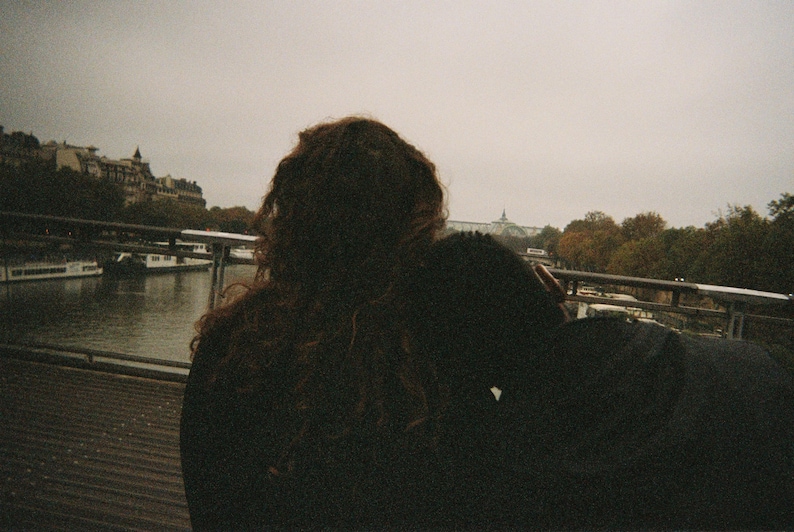 Peut inclure: Photographie d'un moment intime. Deux silhouettes se d&eacute;tachent sur un fond de rivi&egrave;re, de pont et de b&acirc;timents. La sc&egrave;ne est baign&eacute;e de tons sourds, sugg&eacute;rant un jour nuageux. L'accent est mis sur les figures, cr&eacute;ant une sensation de proximit&eacute;.