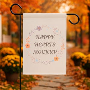 May include: A cream-colored garden flag with the text "HAPPY HEARTS MOCKUP" in gray, surrounded by a floral wreath. The flag is displayed on a black metal stand, with an autumn-themed background.