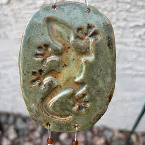 Puede incluir: Un móvil de viento de cerámica hecho a mano con un colgante ovalado verde y marrón. El colgante tiene un diseño en relieve de un gecko. El móvil de viento está ensartado con alambre transparente y cuentas marrones. Aproximadamente 10 cm de largo.