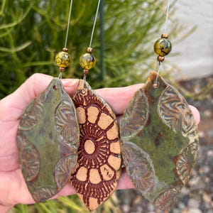 May include: Three ceramic leaf-shaped wind chimes. Two are olive green with embossed leaf patterns, and one is tan with a brown sunburst design. Each leaf hangs from a string with a decorative yellow and brown bead. The background is blurred green foliage.