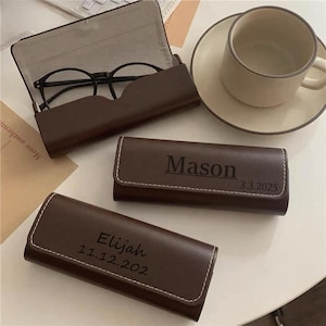 May include: A brown leather glasses case with the name "Mason" and the date "3.3.2025" printed on it. Another case has the name "Elijah" and the date "11.12.202". A pair of black glasses is in an open case. A cup and saucer are in the background.