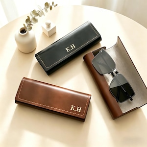 Puede incluir: Tres estuches de cuero para gafas en negro y marrón, con costuras blancas e iniciales doradas. Un estuche está abierto, mostrando gafas de sol negras. Los estuches están sobre una superficie clara.