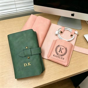 Op de afbeelding: Een verzameling gepersonaliseerde lederwaren in groen en roze tinten. Het groene item is een notitieboek met een gespsluiting en gouden initialen. Een roze item heeft een naam en initiaal. Een witte oplaadkabel is ook zichtbaar.