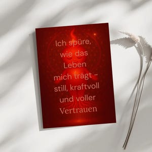 May include: A red card with the text "Ich spüre, wie das Leben mich trägt - still, kraftvoll und voller Vertrauen." A stylized illustration of a figure with a flame-like head is in the center. The card is on a white surface.