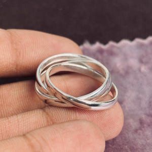 May include: A silver ring featuring three intertwined bands. The bands have a polished finish, reflecting light. The ring is held in a hand, with a soft purple backdrop.