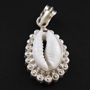 May include: A silver pendant featuring a central white cowrie shell. The shell is surrounded by a decorative border of small, raised silver spheres. A silver bail is attached to the top of the oval-shaped setting, allowing it to be worn on a chain.