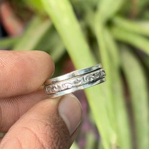 以下が含まれることがあります： 中央に回転するバンドを備えた銀色の金属製リング。バンドには複雑な浮き彫りのスクロール模様が施されています。リングの上下の縁は滑らかに磨かれており、中央の精巧なデザインと対照的な仕上がりになっています。