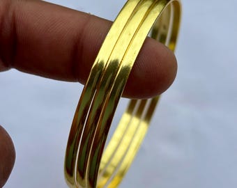 May include: Three gold-colored bangles are held by a person's fingers. The bangles are rectangular in shape and have a smooth, reflective surface. The jewelry is a bright, shiny gold color.