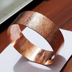 Puede incluir: Un brazalete de color cobre con un diseño floral en relieve. La banda ancha tiene un interior pulido y un exterior texturizado. El brazalete se muestra sobre una superficie blanca con un fondo de madera.