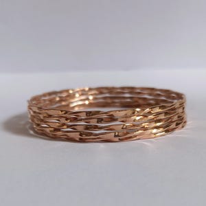Può includere: Un gruppo di cinque braccialetti in oro rosa. Ogni braccialetto presenta un design strutturato e ritorto. I braccialetti sono impilati su una superficie bianca, creando riflessi.