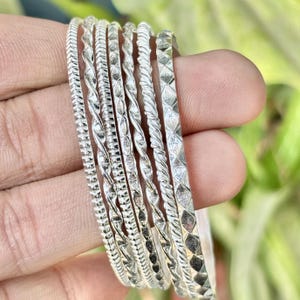 Könnte beinhalten: Eine Sammlung silberner Armreifen mit verschiedenen strukturierten Designs. Die Armreifen weisen Muster wie gedrehtes Seil, Rautenformen und lineare Rillen auf. Der Schmuck wird vor einem verschwommenen grünen Hintergrund präsentiert.