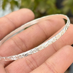 Peut inclure: Un bracelet jonc en argent avec un motif floral. Le bracelet est rond et présente une surface texturée avec des motifs floraux détaillés. Le métal a une qualité brillante et réfléchissante.