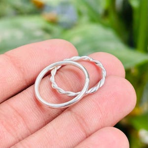 May include: Two silver rings, one with a smooth band and the other with a twisted rope design. The rings are displayed on a person's hand, with a blurred green background.