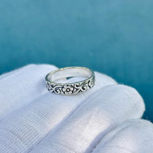 Könnte beinhalten: Ein silberner Ring mit floralem Muster. Der Ring hat ein sich wiederholendes Design aus Blumen und wirbelnden Ranken um das Band. Der Ring wird auf einer weißen Oberfläche vor blauem Hintergrund gezeigt.