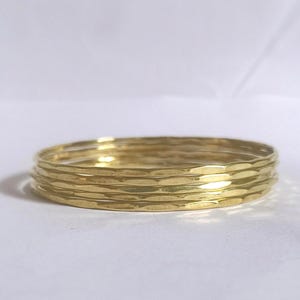 May include: A stack of five gold-colored bangles. Each bangle has a textured, hammered appearance. The bangles are stacked together, creating a layered effect. The background is a plain white surface.