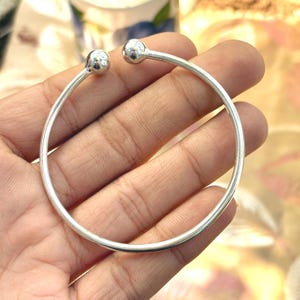 Op de afbeelding: Een zilverkleurige open armband met een gladde, dunne cirkelvormige band die aan beide uiteinden is afgewerkt met een gepolijst, bolvormig accent. Het metaal heeft een reflecterende afwerking en een eenvoudig, doorlopend ontwerp.
