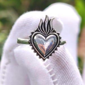 Può includere: Anello argentato con un design a forma di cuore e un motivo a fiamma nella parte superiore. Il cuore è circondato da un bordo smerlato. L'anello è mostrato su uno sfondo bianco.