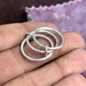 May include: A silver ring featuring three interconnected circles. The rings are joined at two points, forming a distinctive, contemporary design. The ring is held in a hand, with a blurred background.