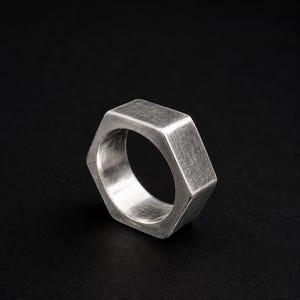 May include: A silver-colored metal ring shaped like a hexagonal nut. The band features a flat, six-sided exterior surface with a brushed metallic finish and a smooth, circular interior opening, presenting a solid and industrial appearance.