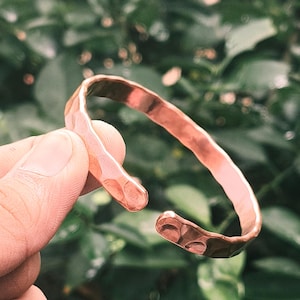 Puede incluir: Un brazalete de puño abierto de color cobre con una textura martillada. El brazalete se sostiene en una mano contra un fondo verde borroso. El metal tiene un tono marrón rojizo cálido.