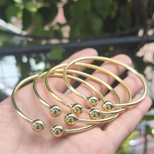 May include: A collection of gold-coloured bangles. Each bangle features a smooth, rounded band with a small sphere at each end. The bangles are presented against a blurred green backdrop.