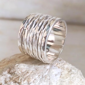 Peut inclure: Une large bague en argent texturé. La bague présente une série de crêtes horizontales en forme de vagues qui entourent le bracelet. L'argent poli reflète la lumière, créant des reflets et des ombres. La bague est posée sur un fond neutre.