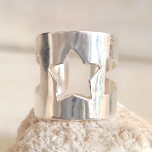 May include: A silver ring featuring a star-shaped cut-out. The ring has a wide band design with a star-shaped void in the middle. The ring is made of a shiny metal and is shown on a textured surface.