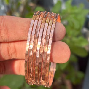 Puede incluir: Un conjunto de pulseras de cobre martillado. Las pulseras tienen una superficie texturizada y un color marrón rojizo cálido. Las pulseras están apiladas, mostrando su diseño y artesanía. El fondo está borroso, con follaje verde visible.