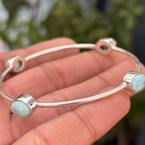 Könnte beinhalten: Ein silbernes Armreif-Armband mit vier ovalen, hellblauen Edelsteinen. Die Edelsteine sind in einzelnen Silberfassungen gefasst. Das Armband wird vor einem verschwommenen grünen Hintergrund gezeigt. Durchmesser: ca. 6 cm.