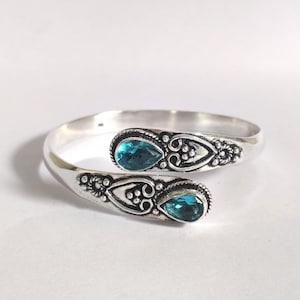 Puede incluir: Un brazalete plateado con detalles ornamentados y dos gemas en forma de lágrima de color verde azulado. El brazalete presenta intrincados patrones alrededor de las gemas y a lo largo de la banda, creando un diseño elegante y llamativo.