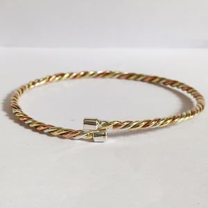 Puede incluir: Un brazalete de metal trenzado con tonos dorados, plateados y cobrizos. El brazalete tiene un diseño sencillo y abierto con un cierre de broche. El metal está trenzado para crear una textura similar a una cuerda. Joyería.