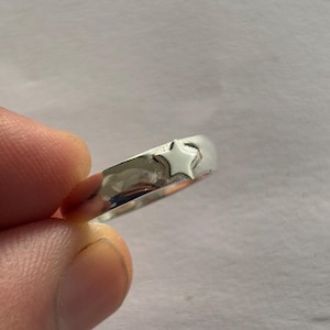 Puede incluir: Un anillo plateado con un diseño de estrella. El anillo es liso y pulido, con una pequeña estrella en relieve en la banda. El anillo está sostenido por los dedos de una persona.