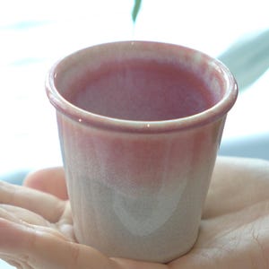 May include: A small, ceramic cup with a pink and white gradient. The cup is held in a hand, showcasing its size and design. The cup's rim is a darker pink, fading to a lighter pink and white at the base. The cup is likely for drinking.
