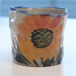 May include: A small, handmade ceramic cup with a rough, textured exterior. The cup features a vibrant, hand-painted sunflower design in shades of yellow, orange, and green. The rim is outlined with a blue band. The cup is sitting on a white surface.