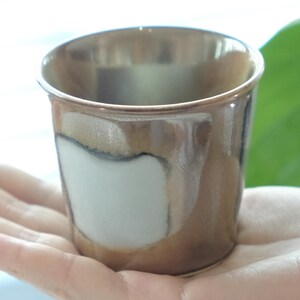 以下が含まれることがあります： メタリックゴールドの縁と茶色と白の釉薬が施された小さな手作りのセラミックカップ。カップは人の手で持たれており、そのユニークなデザインと質感を際立たせています。飲用に使用される可能性が高いです。