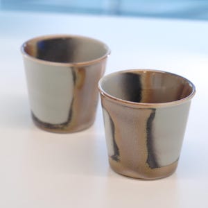 May include: Two ceramic cups with a neutral color palette. The cups have a light gray base with brown and black accents. The cups are cylindrical and appear to be handmade. The background is a white surface with a window.
