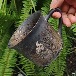 May include: A dark blue ceramic mug with a speckled texture and a black handle. The interior has a golden hue. The mug features an Asian-inspired design and text. The mug is held against a backdrop of green ferns.